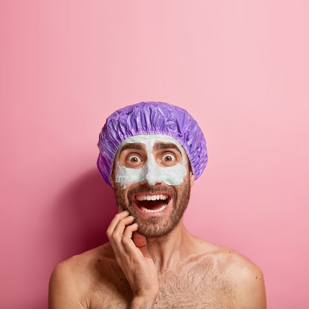 Worried young man applies white mud mask on face, feels refreshment and rejuvenation, wears bathcap, stands shirtless against pink wall