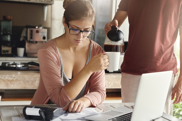 Foto gratuita giovane donna preoccupata che calcola le spese familiari e che fa il bilancio domestico facendo uso del computer portatile e del calcolatore generici in cucina mentre suo marito che sta accanto a lei e che versa il caffè caldo nella sua tazza