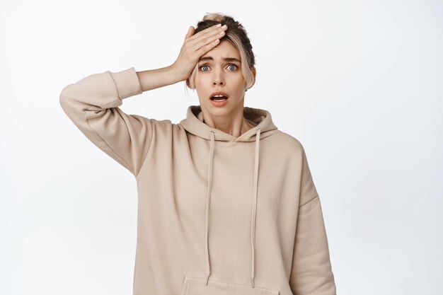 Worried young woman looking alarmed at camera hold hand on head forgot remember something panicking standing anxious against white background