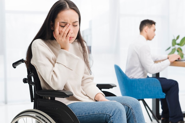 Worried young woman sitting on wheel chair sitting in front of male colleague using laptop