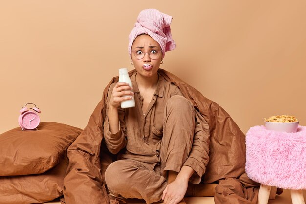 心配している若い女性は頭に包まれたタオルを着て、パジャマはボトルから新鮮なミルクを飲みます茶色の背景の上に分離された毛布の下のベッドで簡単な朝食のポーズをとる朝食時間の概念