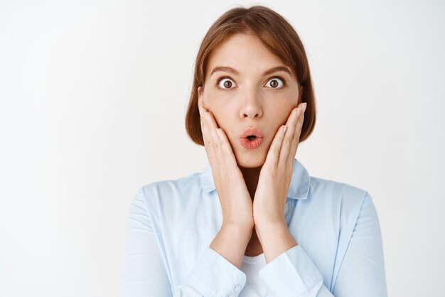 Wow amazing Close up portrait of impressed and surprised young woman look at camera excited with opened mouth holding hands on cheeks checking out promo white background