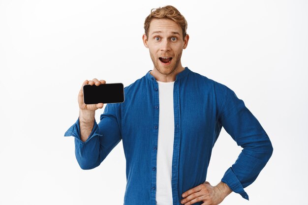 Wow amazing. Young handsome man shows smartphone empty screen and look excited, recommending download app, click on link, standing in casual clothes against white background