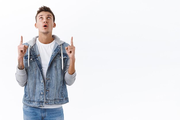 Wow awesome deal look Portrait of speechless amazed blond male queer guy in denim jacket drop jaw in awe looking and pointing fingers up at top advertisement white background