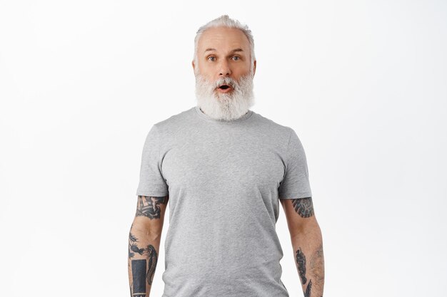Wow awesome. impressed mature man with tattoos and beard, drop jaw, gasping and looking in awe and excitement at special promo offer, shopping deal, standing over white background.