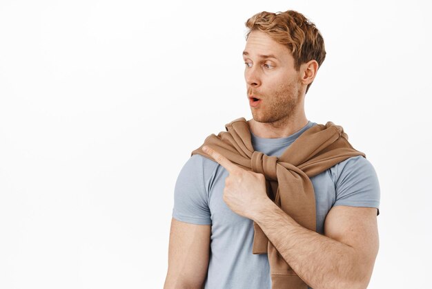 Wow awesome Impressed redhead man gasping pointing finger left and turn head at advertisement showing announcement standing against white background