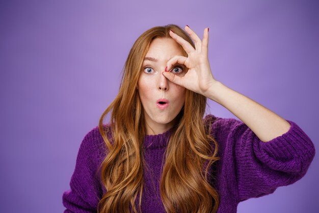 Wow awesome, take closer look. Surprised and impressed attractive redhead female student holding okay or circle sigh over eye and looking through, folding lips in amazement over purple wall.