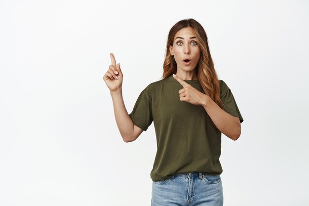 Wow check out. Impressed middle-aged woman, female customer pointing fingers at upper left corner with excited face expression, showing advertisement, white background.