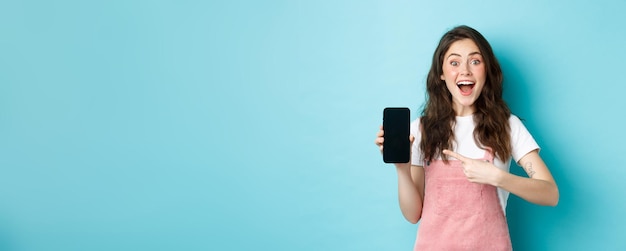 Free photo wow check this out excited pretty girl pointing finger at phone screen showing logo or store advertisement on smartphone standing against blue background