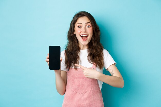 와우 이것을 확인하십시오. 파란 배경에 서서 스마트폰에 로고나 매장 광고를 보여주는 흥분된 예쁜 소녀가 전화 화면을 손가락으로 가리키고 있습니다.