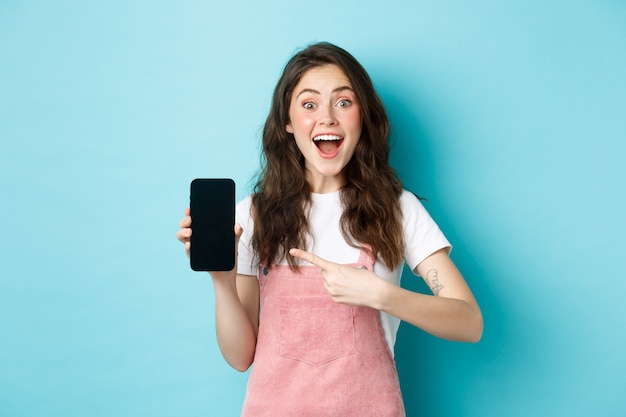 Wow check this out. Excited pretty girl pointing finger at phone screen, showing logo or store advertisement on smartphone, standing against blue background.