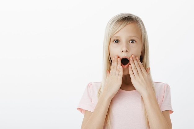 Wow, kid shocked hearing amazing news about favorite cartoon. Indoor shot of amazed and surprised happy blond daughter, gasping, standing with hands near opened mouth and popping eyes, being stunned