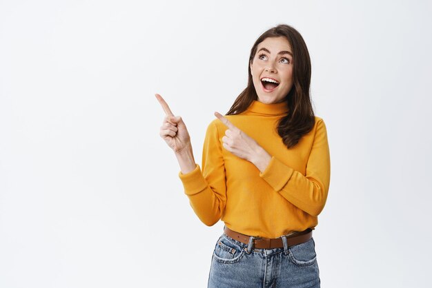 Wow look Excited smiling girl pointing and staring at upper left corner advertising showing awesome promo deal standing amazed against white background