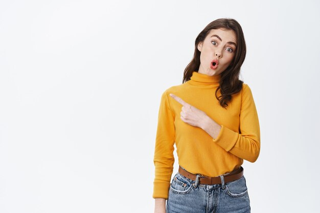 Wow look here Excited brunette girl looking amazed drop jaw and pointing finger left at promo offer standing against white background