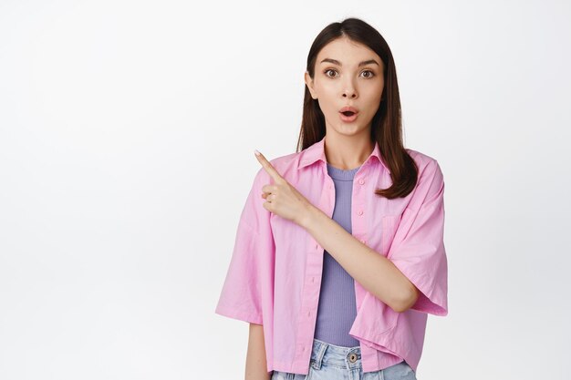 Wow look at that Surprised girl pointing finger left and looking curious asking question about sale banner staring intrigued standing over white background