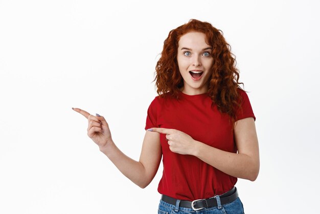 와우 보세요 곱슬머리를 한 흥분한 빨간 머리 유럽 여성이 헐떡이고 프로모션 광고에서 손가락을 가리키며 멋진 제안 흰색 배경을 확인합니다