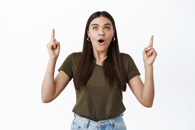 Wow look there Impressed and happy young woman pointing fingers up gasping fascinated and staring at banner logo showing amazing news white background