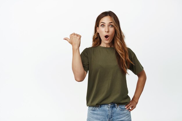 Wow look there. Impressed, surprised adult woman tell big news, pointing thumb left, say wow and stare amazed at camera, checking out advertisement, white background