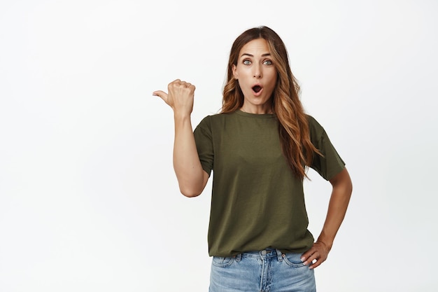 Wow look there. Impressed, surprised adult woman tell big news, pointing thumb left, say wow and stare amazed at camera, checking out advertisement, white background
