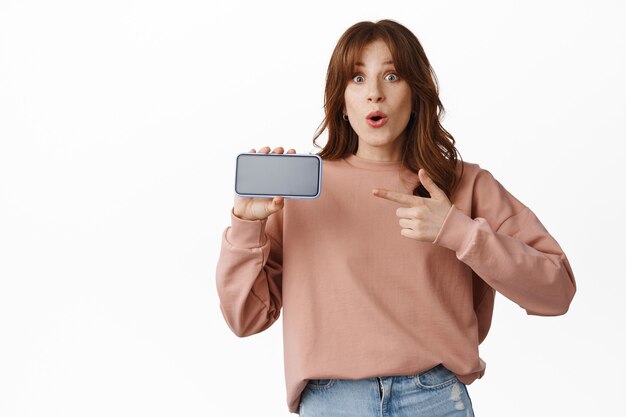 うわー、このアプリを見てください。興奮した笑顔の生姜の女の子がスマートフォンの水平画面に指を指して、携帯電話でアプリを表示し、白の上に立っています。