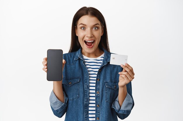 Wow look at this online sale. Excited woman showing her smartphone screen, mobile app interface and credit card, smiling amazed, standing over white background.