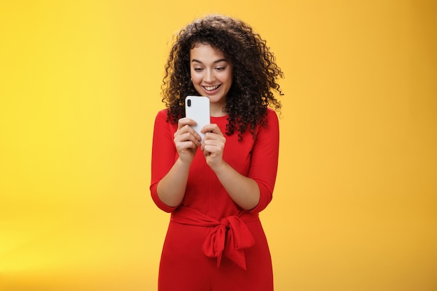 Ничего себе новый мобильный телефон потрясающий. Впечатленная и изумленная симпатичная кудрявая женщина в красном платье, держащая смартфон, смотрящая на экран, забавляется, играя в крутое приложение или игру через желтую стену.