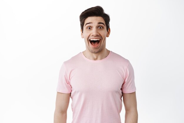 Wow so awesome. Excited handsome man drop jaw from amazement, smiling and look fascinated, checking out cool promo deal, stare at advertisement, standing over white background