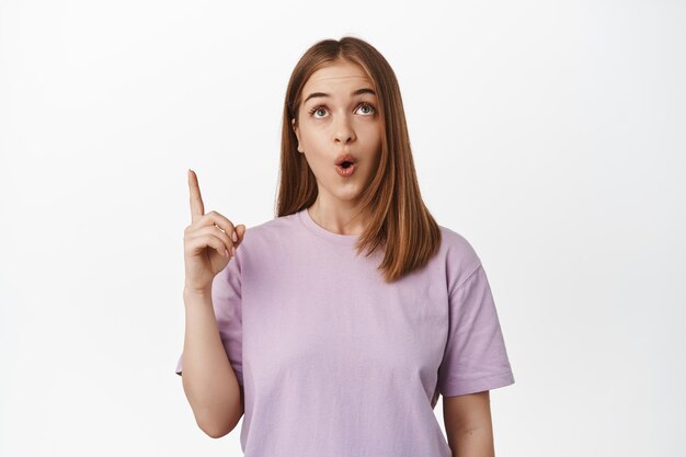 Wow so awesome. Young woman pointing and looking up with fascinated, impressed face, found smth in store worth attention, standing over white background in summer t-shirt