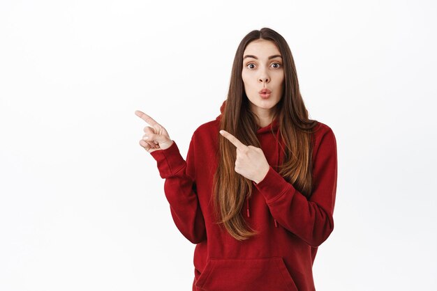 Wow thats interesting have you seen this Curious girl look intrigued while pointing fingers aside showing promo deal on left copy space standing in red hoodie against white background