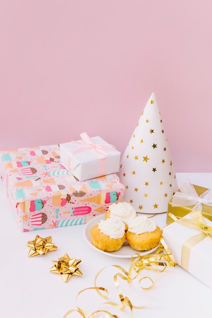 Wrapped gift boxes; bow; streamer; party hat and cupcakes on desk against pink background