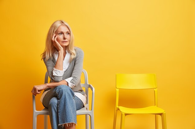 wrinkled blonde European woman being deep in thoughts sits on comfortable chair waits for something feels lonely and melancholic wears stylish clothes