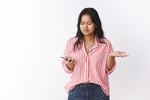 Wtf it supposed mean. Portrait of confused and frustrated young girlfriend standing questioned shrugging and frowning as holding smartphone reading strange message in mobile phone over white wall