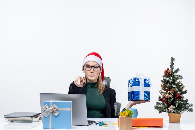 Umore festivo di natale con la giovane donna positiva curiosa con il cappello di babbo natale e gli occhiali da portare che si siedono ad un tavolo che mostra il regalo che indica qualcuno su fondo bianco