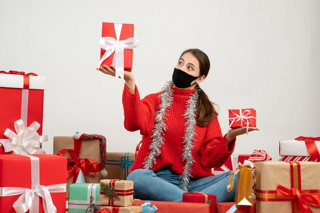 xmas girl with black mask holding gifts in both hands sitting around presents on white