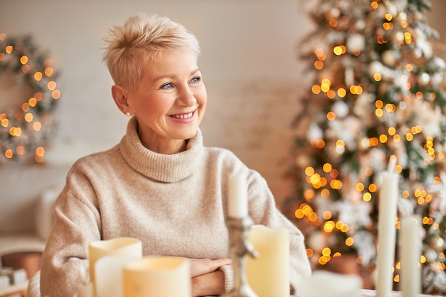 無料写真 クリスマス、休日、装飾、パーティー、お祭りの雰囲気のコンセプト。クリスマス気分を楽しんで、ワックスキャンドル、装飾、ライトの周りに座って、短い髪の格好良い陽気な中年女性