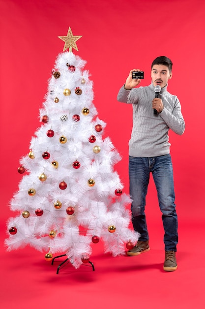 Umore natalizio con ragazzo fiducioso vestito in jeans in piedi vicino all'albero di natale decorato e tenendo il microfono per scattare la sua foto