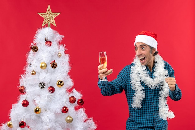 クリスマスツリーの近くでワインのグラスを保持している青いストリップシャツでサンタクロースの帽子をかぶった狂気の感情的な若い男とクリスマスムード