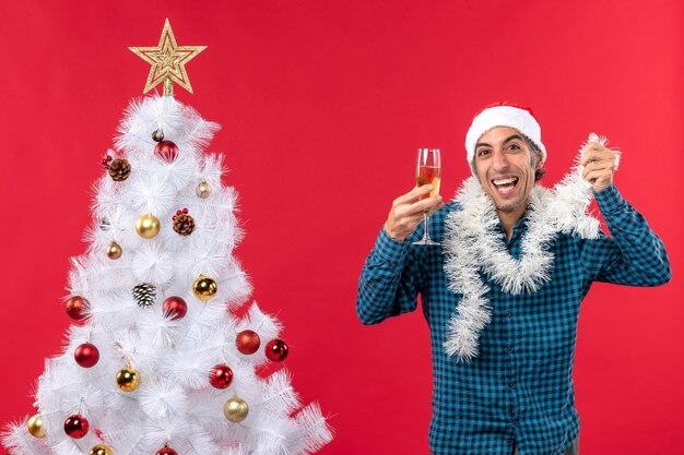 サンタクロースの帽子をかぶって、クリスマスツリーの近くでワインのグラスを上げるクレイジーな感情的な若い男とクリスマスムード