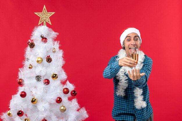 クリスマスツリーの近くでワインのグラスを上げる青い縞模様のシャツにサンタクロースの帽子をかぶった感情的な面白い若い男とクリスマスムード