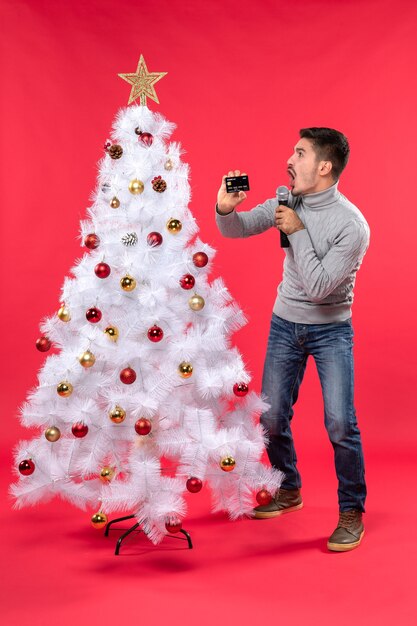 рождественское настроение с эмоциональным парнем, стоящим возле украшенной елки и держащим микрофон и телефон забавными жестами