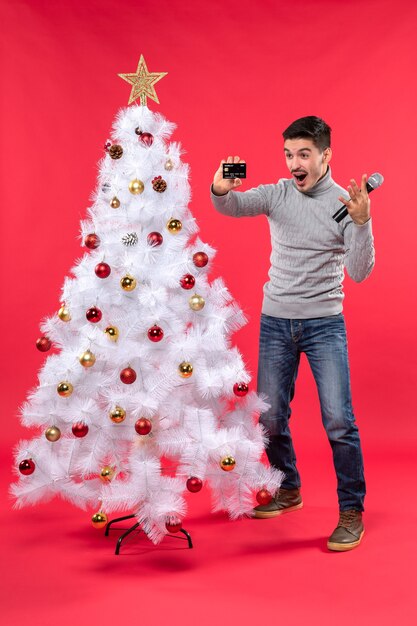 装飾されたクリスマスツリーの近くに立って、マイクと電話を持っている感情的な男とクリスマス気分