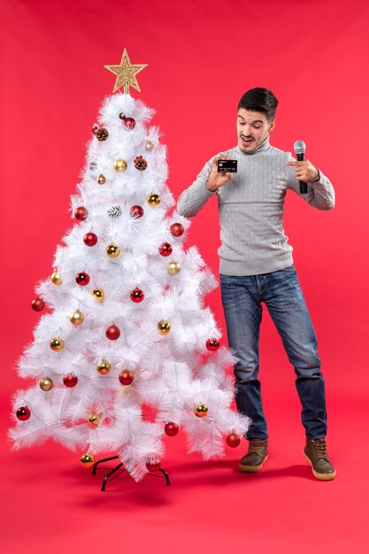 рождественское настроение с эмоциональным парнем, стоящим возле украшенной елки, держащим микрофон и указывающим на телефон