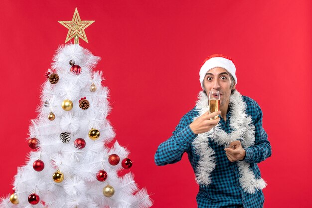 рождественское настроение с эмоциональным молодым человеком в шляпе санта-клауса в синей полосатой рубашке, пьющим бокал вина возле елки