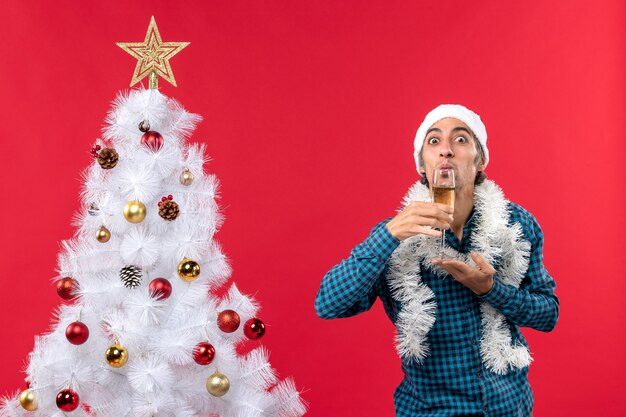 xmas mood with emotional young man with santa claus hat in a blue stripped shirt holding and drinking a glass of wine near Christmas tree