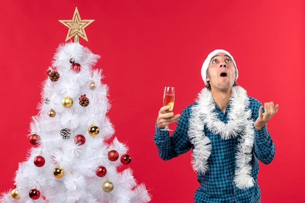 рождественское настроение с эмоциональным молодым человеком в шляпе санта-клауса в синей полосатой рубашке, держащим бокал вина и смотрящим вверх возле елки
