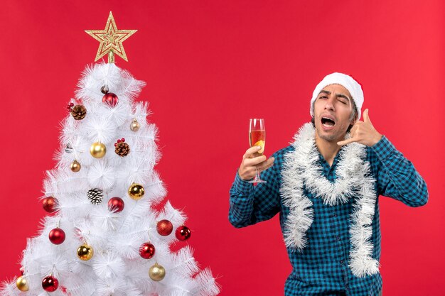 xmas mood with emotional young man with santa claus hat in a blue stripped shirt holding a glass of wine and making call me gesture near Christmas tree