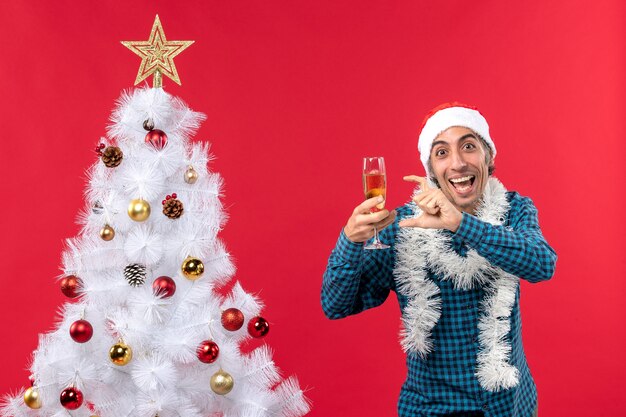 クリスマスツリーの近くに何かを指しているワインのグラスを保持している青い縞模様のシャツにサンタクロースの帽子をかぶった感情的な若い男とクリスマスムード