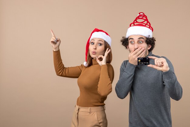 xmas mood with excited cool couple wearing red santa claus hats guy showing bank card. woman pointing above