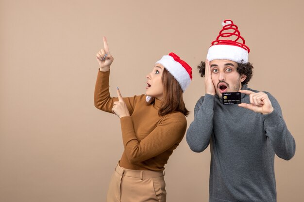 Umore natalizio con coppia cool eccitata che indossa cappelli rossi di babbo natale arrabbiato ragazzo che mostra carta di credito. donna che punta sopra