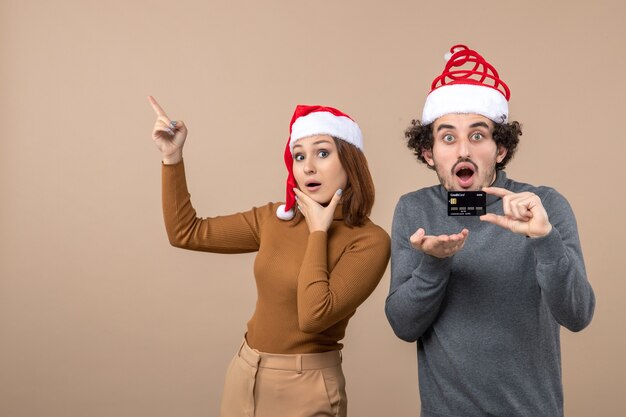 рождественское настроение с возбужденной довольной крутой парой в красных шапках санта-клауса, парень показывает банковскую карту, женщина показывает вверху